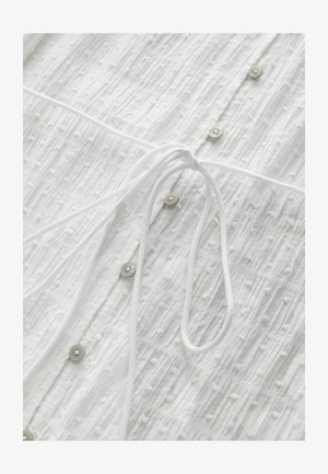 Hvid top med tekstureret mønster, med små knapper og en snøre til at binde. Blødt stof med forhøjede, vekslende striber.