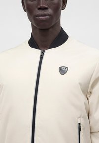 Άνδρας που φοράει ανοιχτό μπεζ jacket με φερμουάρ, μαύρο γιακά και λογότυπο EA7 Emporio Armani στο στήθος.