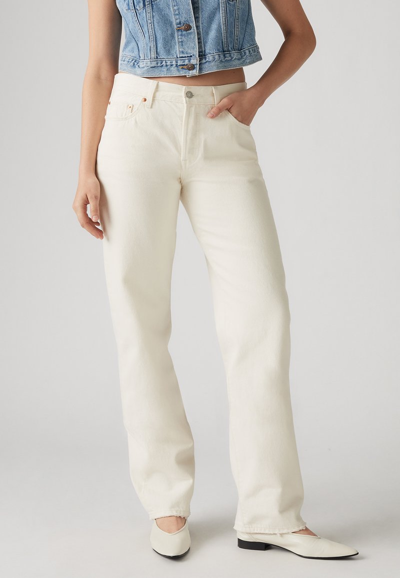 Γυναίκα που φοράει παντελόνι τζιν ίσιο, εκρού, με ψηλή μέση, λευκές μυτερές φλατ, και ένα κοντό, χωρίς μανίκια, τζιν μπουφάν, στέκεται με ένα χέρι στην τσέπη.