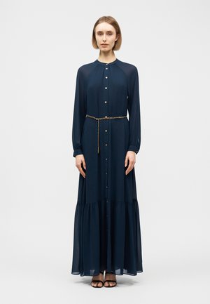 Mujer de pie con un vestido largo azul marino abotonado, cinturón de cadena dorada y tacones negros abiertos al frente, sobre un fondo claro y sencillo.