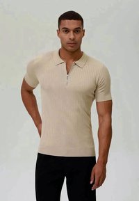 Beige räfflad kortärmad polo med dragkedja i halsen, figurnära design och mjuk textur. Bärs ihop med svarta byxor.