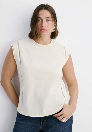 Femme aux cheveux châtains mi-longs porte un haut crème sans manches orné de dentelle, un jean foncé, une bague argentée et des boucles d’oreilles argentées, posant devant un mur blanc.