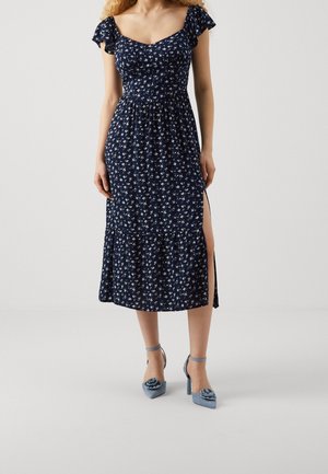 Vrouw draagt een marineblauwe midi-jurk met bloemmotief, wijd uitlopende mouwen en een split aan de zijkant, gecombineerd met blauwe hoge hakken met bloemaccenten.