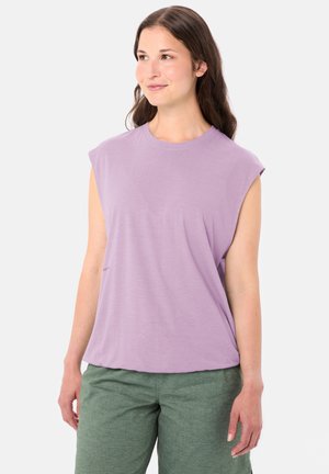 Frau mit langem, dunklem Haar, die ein ärmelloses fliederfarbenes Oberteil und grüne Hose trägt, steht entspannt und schaut leicht zur Seite.