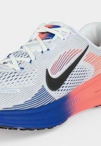 Λευκό δρομικό παπούτσι Nike με μαύρο λογότυπο, μπλε και κοραλλί διαβαθμισμένη σόλα, λευκά κορδόνια και πλεκτή υφή στο επάνω μέρος.