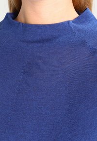 Blå stickad topp med hög halsringning, ribbad textur och subtila sömdetaljer. Tyget verkar mjukt och lätt.