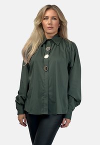Ciemnozielona koszula zapinana na guziki, wykonana z gładkiego materiału, z ostro zakończonym kołnierzykiem i trzema dużymi metalowymi guzikami z przodu.