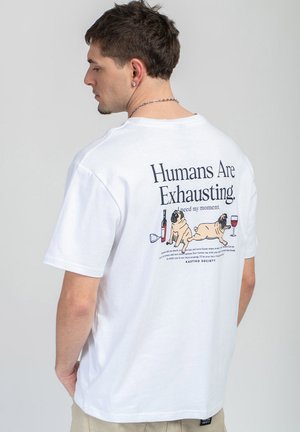 Mann trägt weißes T-Shirt mit dem Text „Menschen sind anstrengend“ und einer Illustration von zwei Möpsen mit Weinflaschen und Gläsern auf dem Rücken.