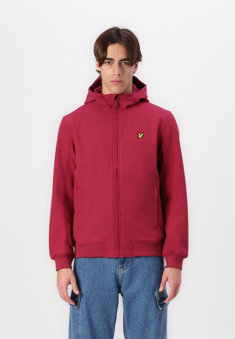 Sweatshirt vermelho com fecho zip, com um acabamento texturizado, apresentando capuz e bolsos laterais, e um pequeno logótipo verde no peito. Combinado com jeans azul claro.