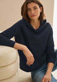 Sudadera azul marino con cuello tipo cowl, hecha de tela suave. Presenta mangas largas y un corte relajado. Combinada con jeans azules desgastados.