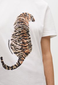Sequin embellishment of a sitting tabby cat in black, brown, and beige on a white short-sleeve shirt with "LOLA CASADEMUNT" text.