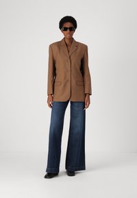 Brun skreddersydd blazer med en enkelt brystlomme, kombinert med vide mørkeblå denimjeans og svarte solbriller.