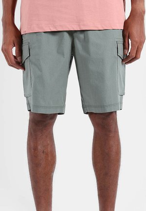 Homme portant un short cargo gris et une chemise rose, debout avec les mains détendues sur les côtés devant un fond uni.