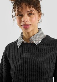 Czarny prążkowany sweter dziany noszony na szarej i białej koszuli w kratę z kołnierzykiem, z okrągłym dekoltem i fakturowaną tkaniną.