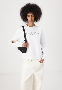 Felpa bianca con collo a giro e logo testuale, polsini elasticizzati e dettagli a taglio. Abbinata a jeans larghi di colore chiaro e una borsa nera.