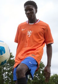 Oranje voetbalshirt met wit Nike-logo, leeuwenkruis en meshpatroon. Blauwe shorts, zwarte sokken, speler in actie met een bal.