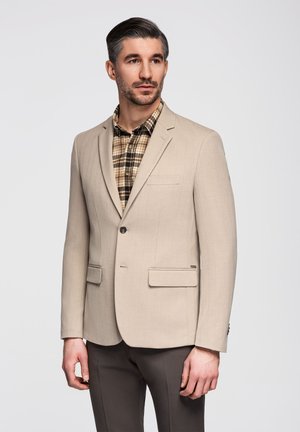 Homme aux cheveux foncés et à la barbe portant un blazer beige sur une chemise à carreaux marron et un pantalon foncé devant un fond clair uni.