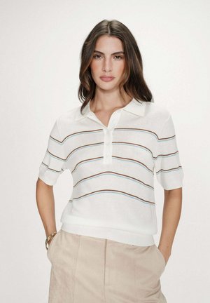 Femme aux longs cheveux foncés portant un polo blanc à manches courtes avec des rayures marron et bleues et un pantalon beige, les mains dans les poches.