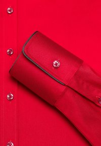 Camicia rossa con una superficie liscia, bottoni lucidi e piping nero a contrasto lungo il polsino, mostrata parzialmente spiegata.