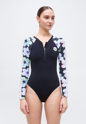 Γυναίκα που φοράει μαύρο μακρυμάνικο μαγιό με πολύχρωμα φλοράλ μανίκια και φερμουάρ στο μπροστινό μέρος, στέκεται μπροστά σε απλό ανοιχτόχρωμο φόντο.