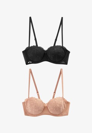 Donna sorridente che indossa un reggiseno nero in pizzo senza spalline e biancheria intima in pizzo abbinata, in piedi contro un muro bianco con i capelli raccolti in uno chignon.