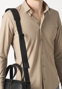 Beige långärmad skjorta med krage, med knappstängning. En svart axelväska med rem bärs, vilket visar släta ytor.