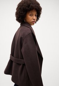 Cappotto avvolgente marrone con ampio colletto, vita sfiancata e spalle strutturate. Tessuto morbido, silhouette aderente e tasche laterali.