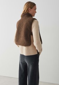 Gilet en polaire marron et duveteux avec un col haut, superposé à un pull crème à côtes et un jean noir à jambe droite. Fond uni.