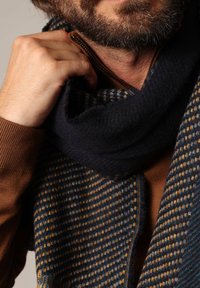 Écharpe en laine bleu marine avec un motif texturé et des accents jaune-orange, drapée sur un pull marron. Gros plan sur le tissu et le détail de la fermeture éclair.