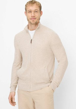 Blonder Mann lächelt, trägt einen beigen Reißverschluss-Pullover über einem weißen Hemd und beigen Hosen, steht mit einer Hand in der Tasche vor weißem Hintergrund.