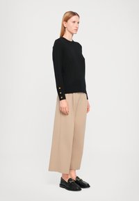 Zwart langarm trui met gouden knoopaccenten op de manchetten, gecombineerd met beige wijde broek en zwarte loafers.