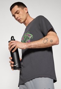 Grå bomulds t-shirt med et farverigt grafisk design, parret med en sort vandflaske i rustfrit stål med Nike-logoet.