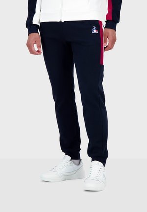 Pantalon de jogging marine en tissu doux avec des accents rouges sur les côtés ; dispose de poignets élastiques et d'un logo brodé sur la cuisse gauche. Porté avec des baskets blanches.