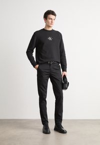 Sweatshirt noir avec logo sur la poitrine, associé à un pantalon noir et des chaussures noires. Design simple, couleurs unies et style ajusté.