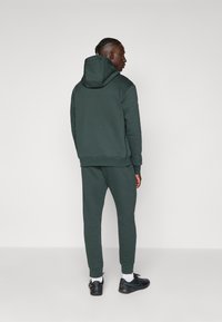 Mørk grønn hettegenser med hette, myk tekstur og elastiske mansjetter; kombinert med matchende joggebukser og svarte treningssko.