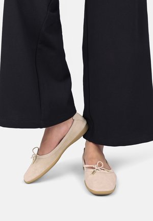 Beige balletsko med små sløjfer båret med sorte bukser med vide ben, vist fra midt på læggen til fødderne.