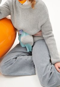 Suéter gris acanalado de manga larga sobre una bufanda de rayas en tonos pastel. Pantalones de mezclilla azul claro; objeto inflable naranja a la izquierda.