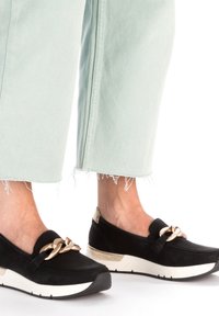Scarpe slip-on nere in suede con un accento a catena, presentano una suola intermedia bianca e dettagli dorati sul tallone. Indossate con pantaloni cropped verde chiaro.