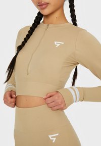 Langärmliges, mit Reißverschluss versehenes Cropped-Top aus beiger Stoffqualität mit weißem Logo und gestreiften Akzenten an den Ärmeln. Es zeichnet sich durch eine taillierte Passform und eine gerippte Textur aus.