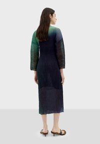 Langes, gradientfarbenes Kleid in dunklen Blau- und Grüntönen, mit plissiertem Stoff und langen Ärmeln, kombiniert mit Sandalen mit pelzigen Absätzen.