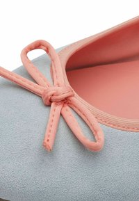 Botte plate en suède gris avec un détail en tissu rose en forme de nœud. Dispose d'un bout arrondi et d'une doublure intérieure lisse. Vue en gros plan du nœud.