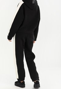 Felpa nera con striscia laterale bianca, dotata di cordoncino e logo sul retro. Abbinata a pantaloni della tuta neri con striscia laterale.