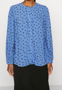 Blusa azul de manga larga con escote redondeado. Presenta pequeños patrones florales oscuros y cierre de botones en la parte delantera. Tejido suave; corte holgado.
