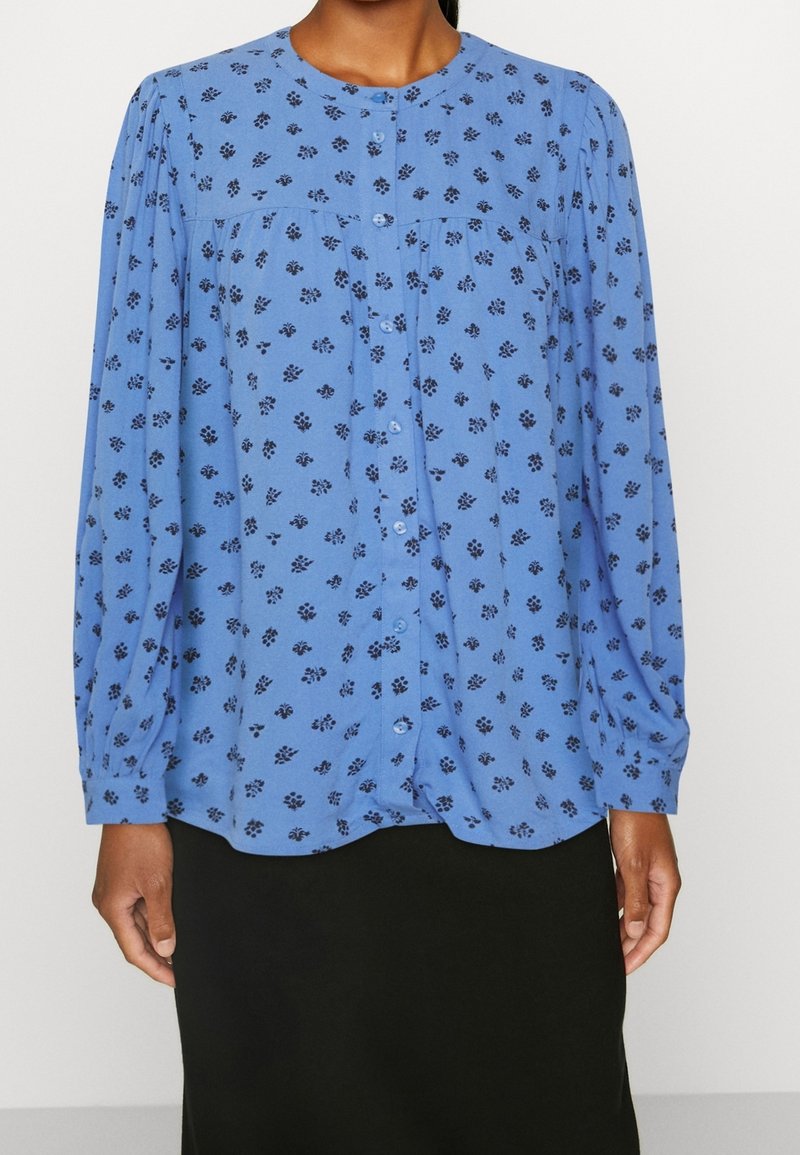 Blusa azul de manga larga con escote redondeado. Presenta pequeños patrones florales oscuros y cierre de botones en la parte delantera. Tejido suave; corte holgado.