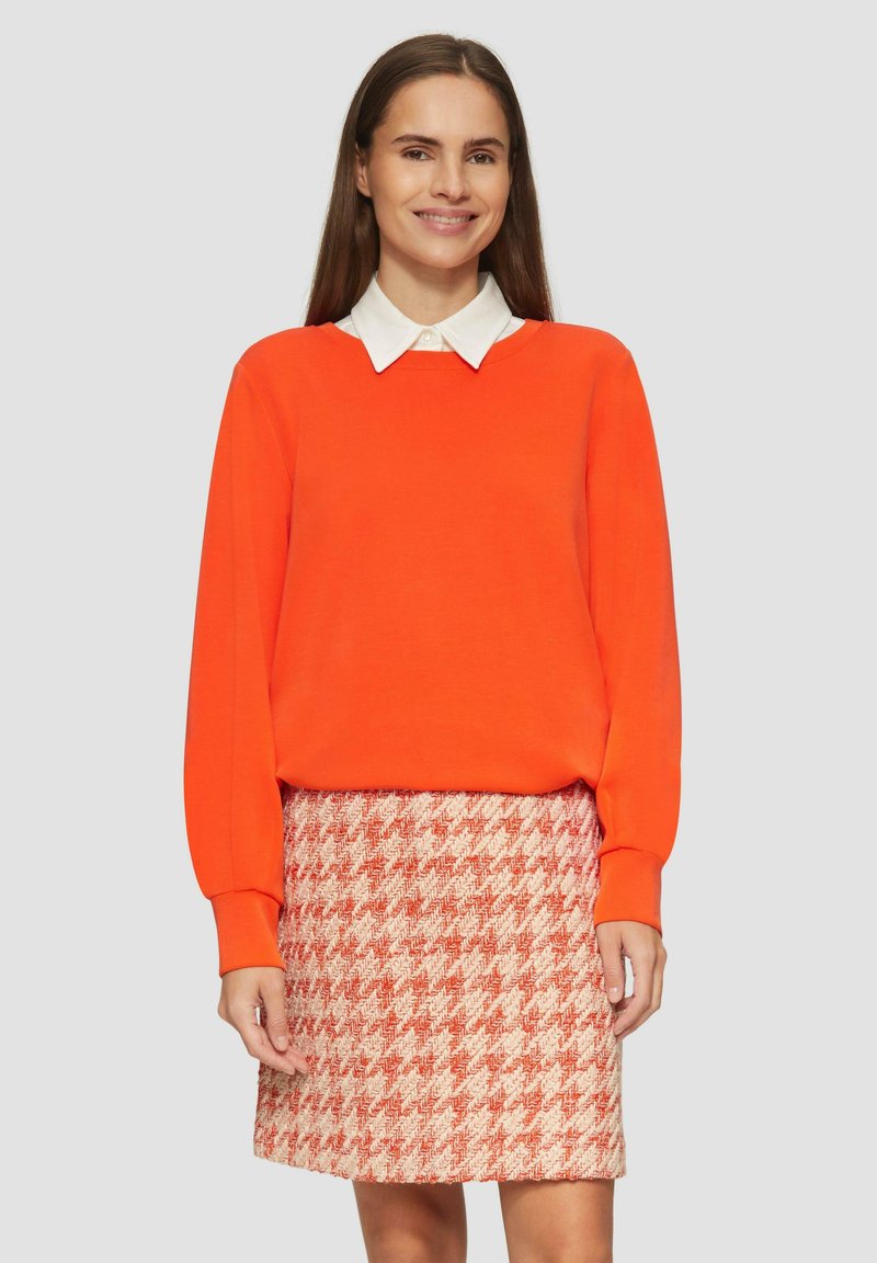 Oranje trui met lange mouwen en een witte coltrui eronder, gecombineerd met een gestructureerde rode hounds-tooth rok.