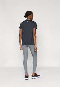 Άτομο με κοτσίδες, που φοράει μαύρο τοπ κοντομάνικο, γκρι κολάν και μαύρα παπούτσια για τρέξιμο, στέκεται στραμμένο με την πλάτη προς τα εμάς σε απλό φόντο.