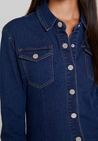 Mörkblå denimskjorta med klassisk krage, två bröstfickor och silverfärgade tryckknappar framtill. Har kontrasterande sömmar.