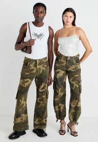 Tarnfarbene Cargo-Hosen mit elastischen Bündchen, tiefen Taschen und verwaschenen Akzenten; kombiniert mit einem weißen Tanktop und einem glitzernden silbernen Neckholder-Oberteil.