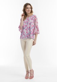 Top floreale con tonalità di rosa e viola, maniche a campana lunghe, abbinato a leggings beige e sandali rosa con cinturini, su uno sfondo bianco.