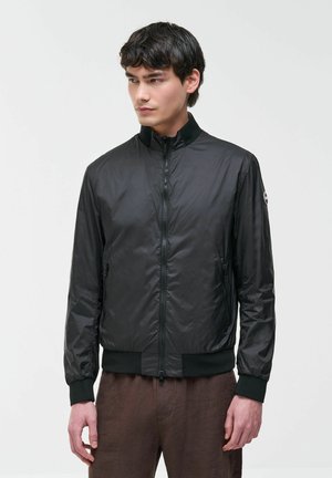 Ung mand iført sort letvægtsjakke med lynlås og brune bukser, stående mod en ensfarvet lys baggrund og kigger mod venstre.